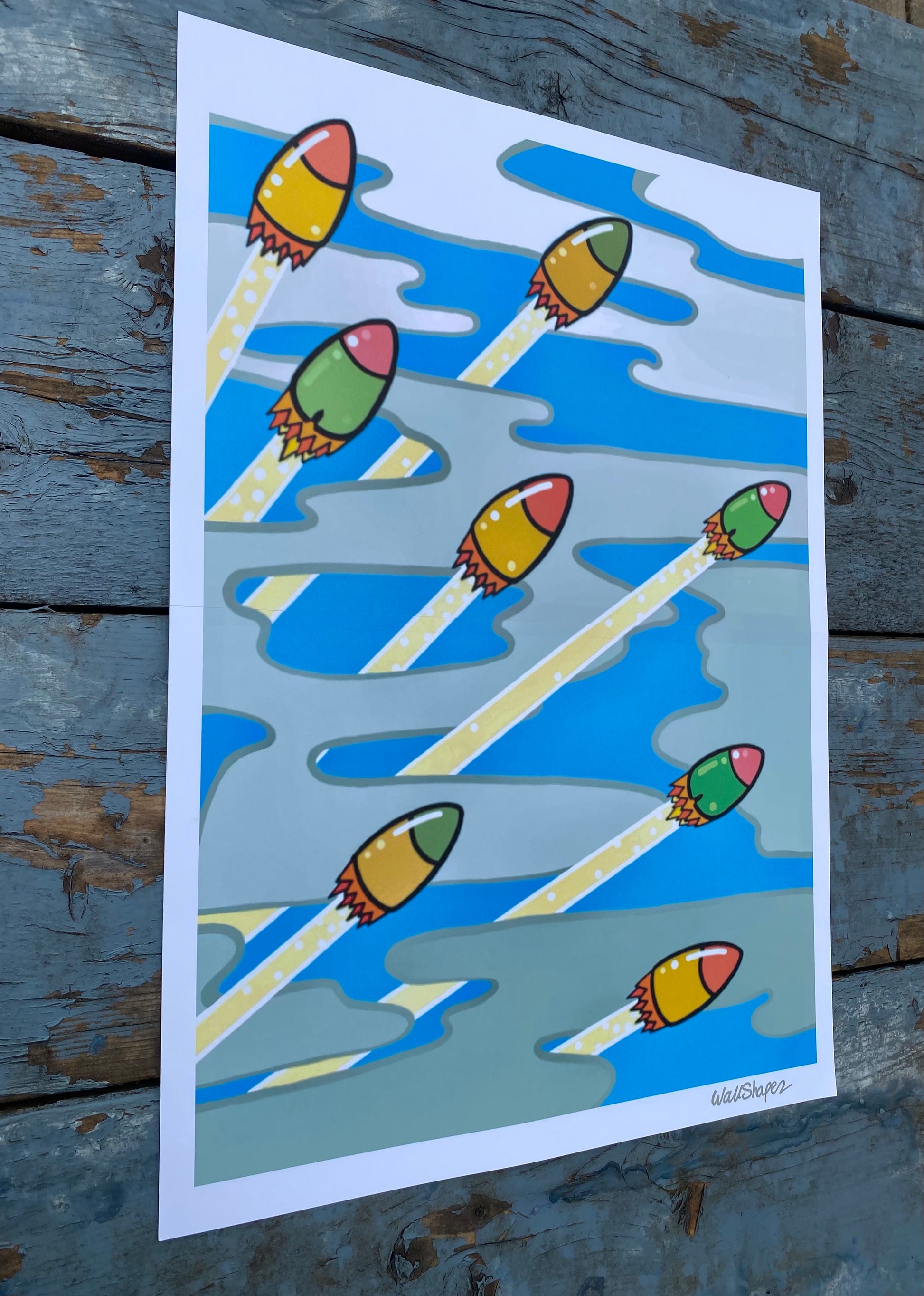 Colorful printed stencil of rockets launching through a cloudy sky, with metallic silver WallShapes signature in the bottom right corner. Photographed in outdoor lighting.