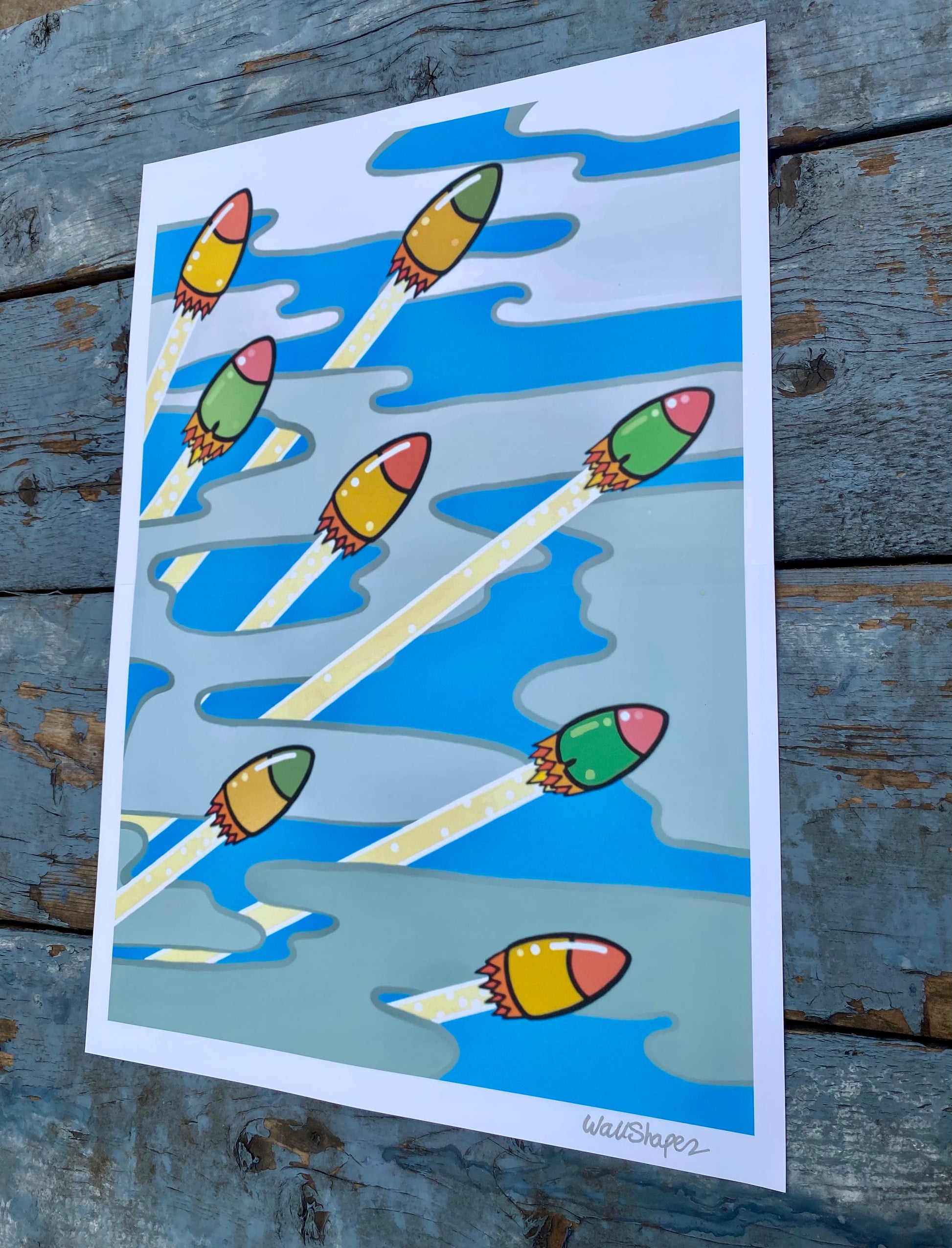 Colorful printed stencil of rockets launching through a cloudy sky, with metallic silver WallShapes signature in the bottom right corner. Photographed in outdoor lighting.