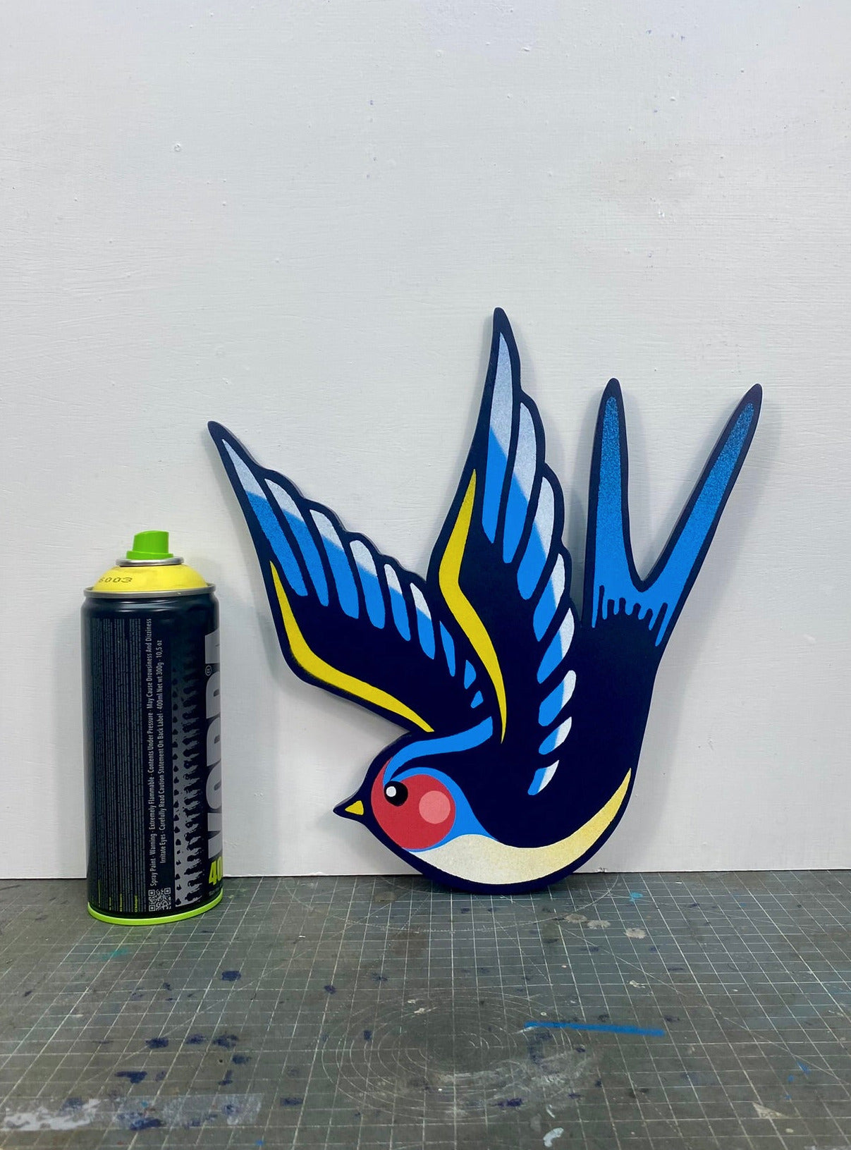 Tattoo inspired, colourful spray painting, painted on a hand-shaped wooden panel displayed on a stencil cutting mat, with a white wall. And a spray can for size comparison.