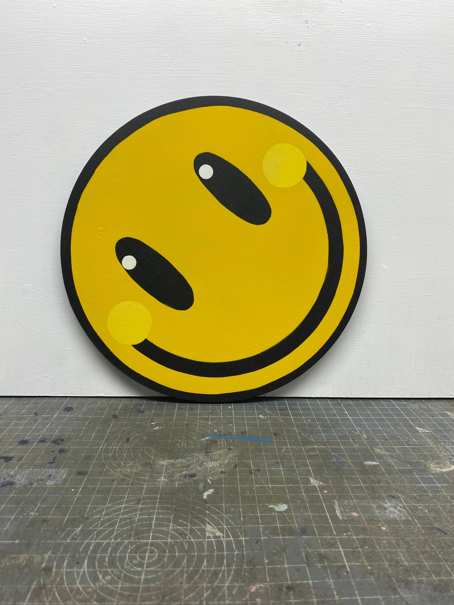 A bright circular yellow smiley face wih yellow cheeks and white eye glints with black boarder on a painted white wall.