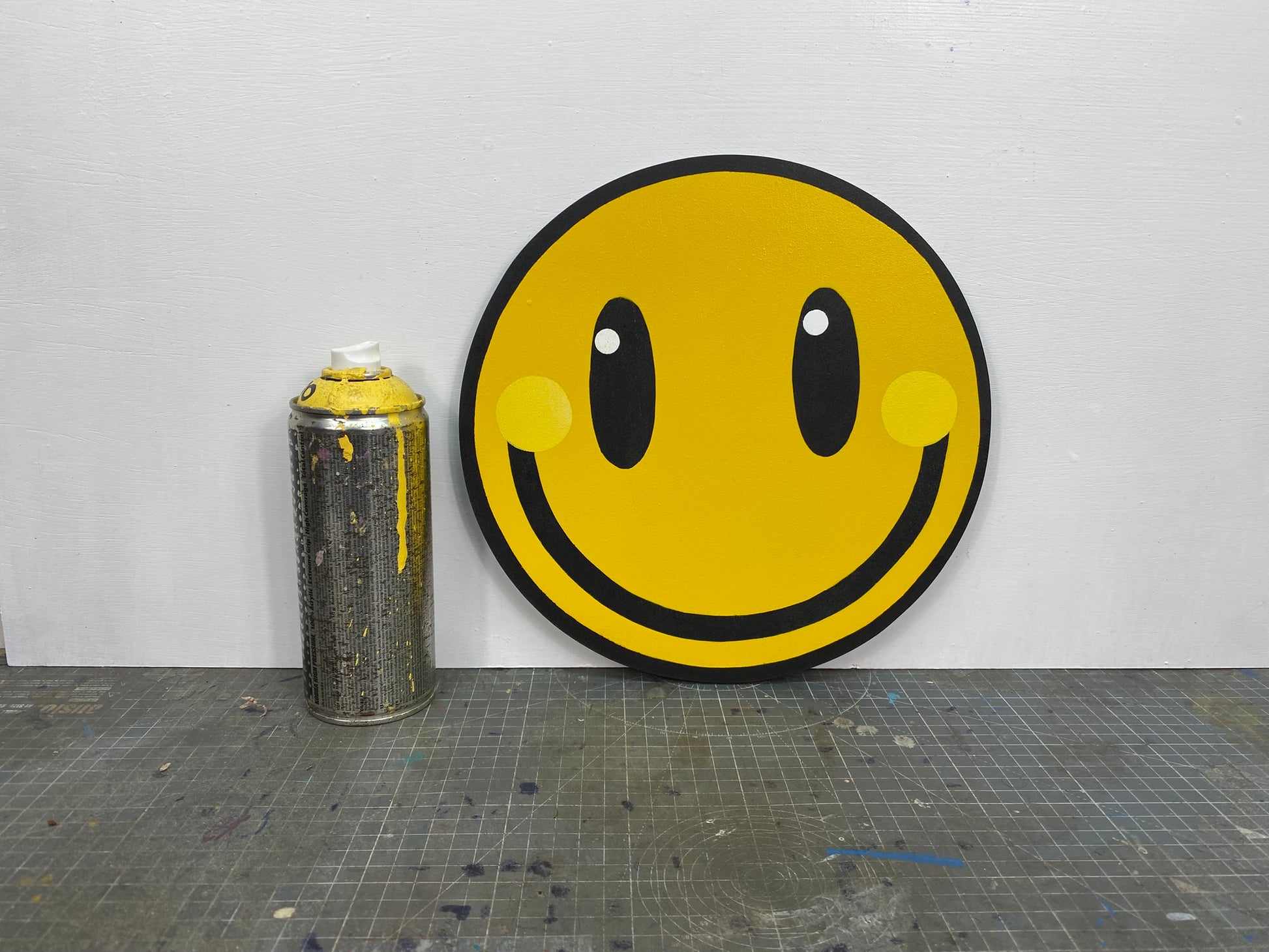 Smiley face design spray painting on a circular panel next to a can of spray paint on a gray cutting mat.