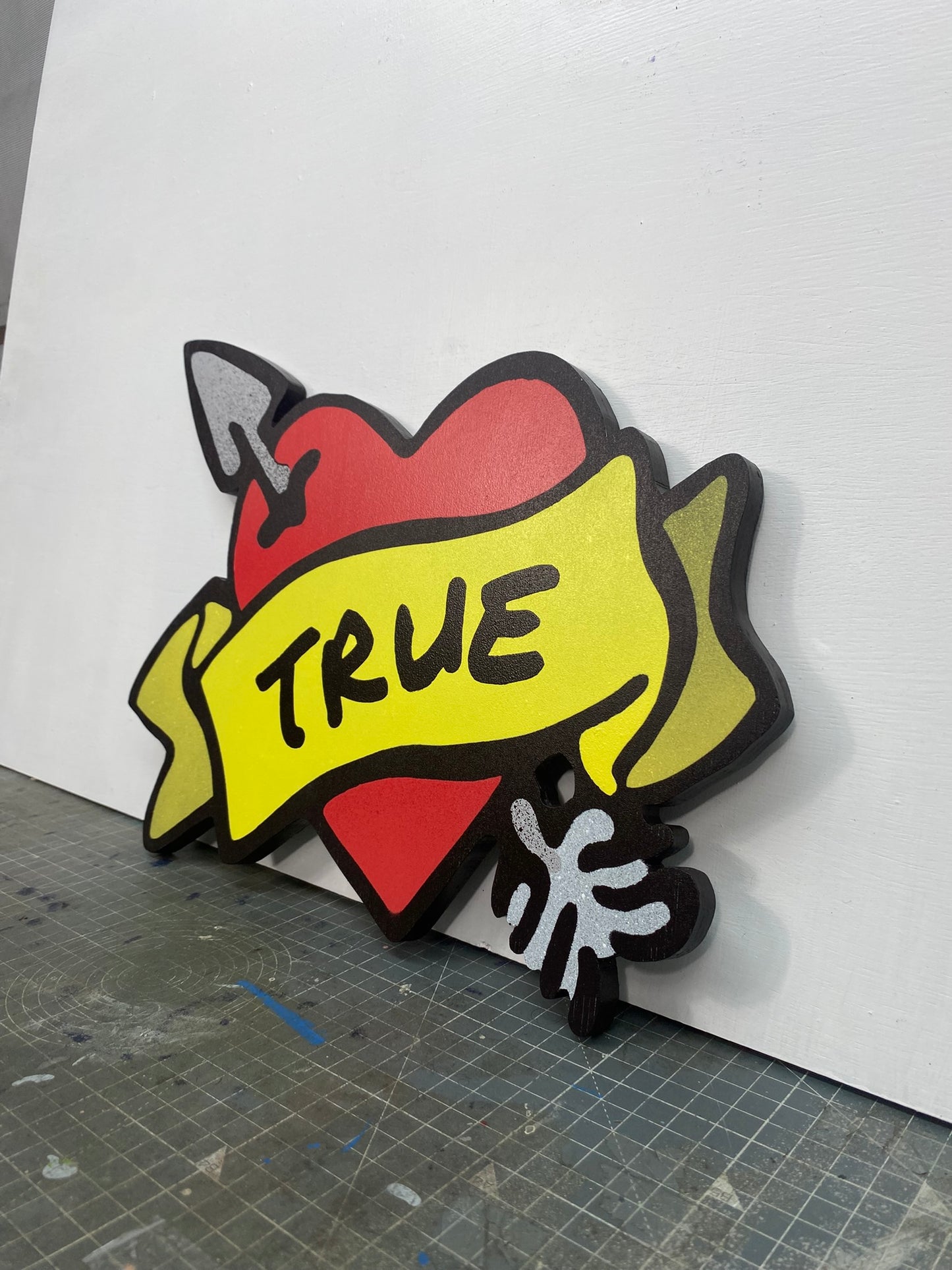 A shaped spray painted art panel of a symbolic pierced love heart with an unfurled banner with TRUE written on it. Photographed in artists studio.