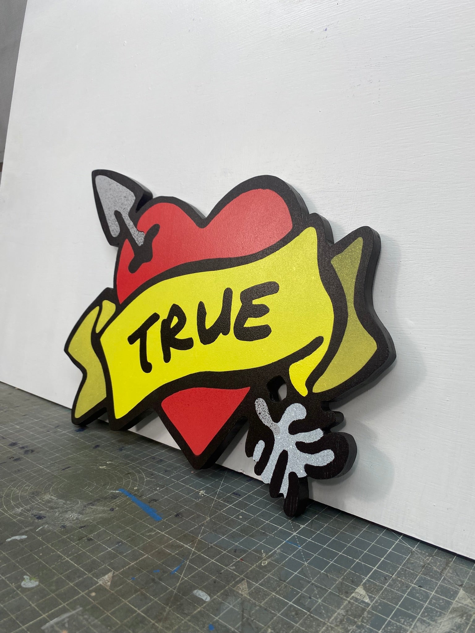 A shaped spray painted art panel of a symbolic pierced love heart with an unfurled banner with TRUE written on it. Photographed in artists studio.