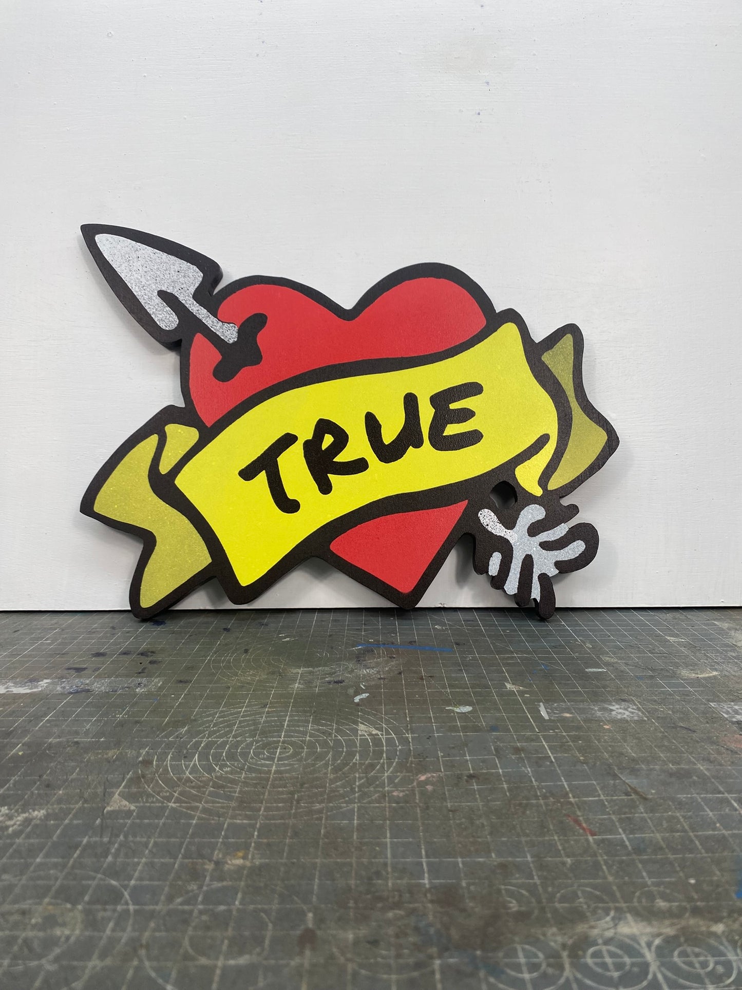 A shaped spray painted art panel of a symbolic pierced love heart with an unfurled banner with TRUE written on it. Photographed in artists studio.