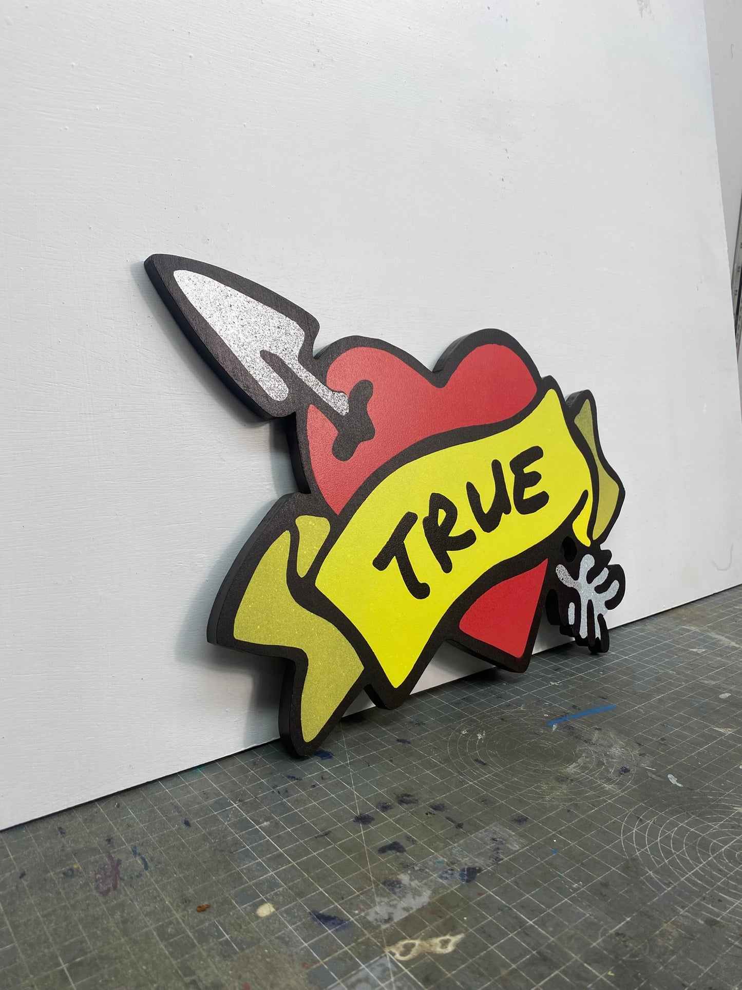 A shaped spray painted art panel of a symbolic pierced love heart with an unfurled banner with TRUE written on it. Photographed in artists studio.