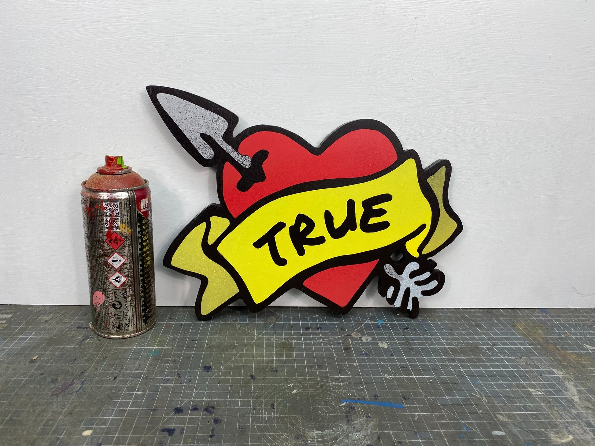 Size comparison of a paint spray can and a shaped spray painted art panel of a symbolic pierced love heart with an unfurled banner with TRUE written on it. Photographed in artists studio.