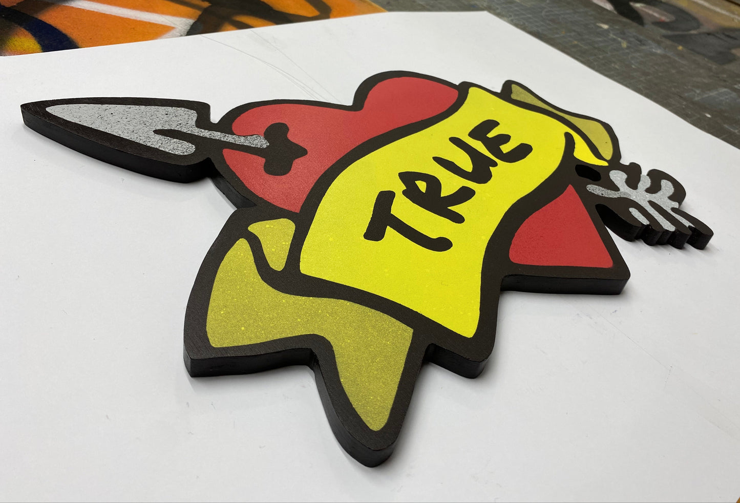 Decorative spray painted, wooden shaped wall art with a red heart, yellow banner, and black TRUE text hanging on a white background.