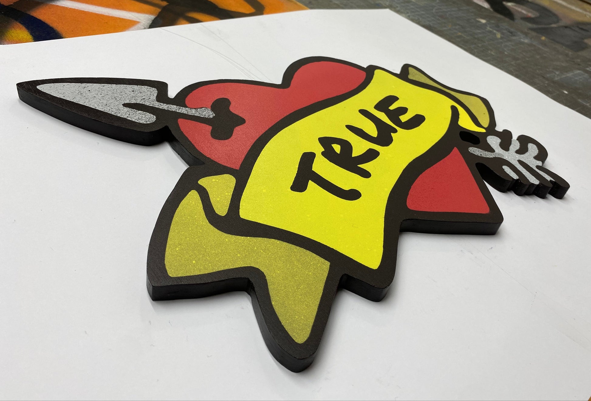 Decorative spray painted, wooden shaped wall art with a red heart, yellow banner, and black TRUE text hanging on a white background.