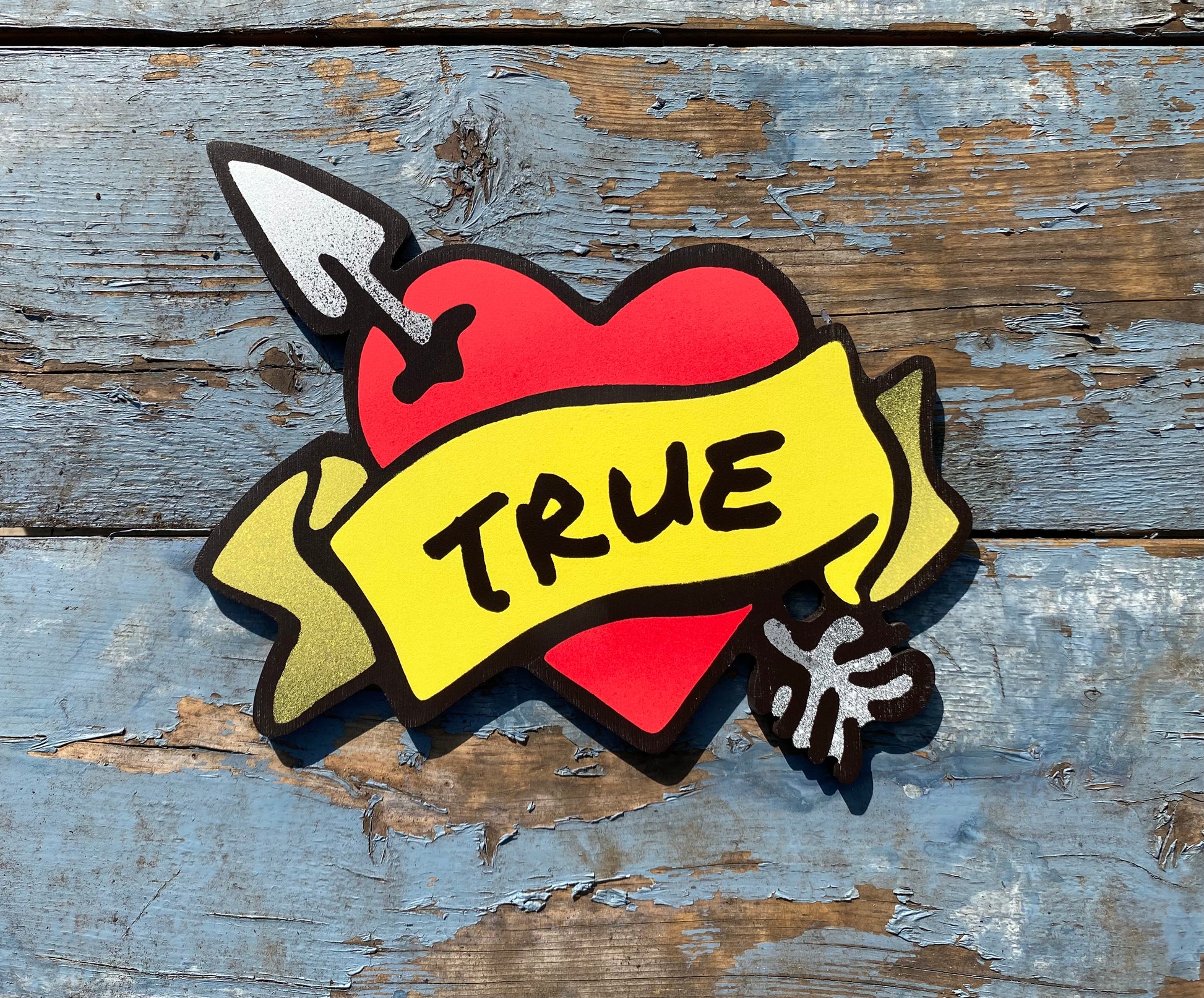 Decorative spray painted, wooden shaped wall art with a red heart, yellow banner, and black TRUE text hanging on a white background.