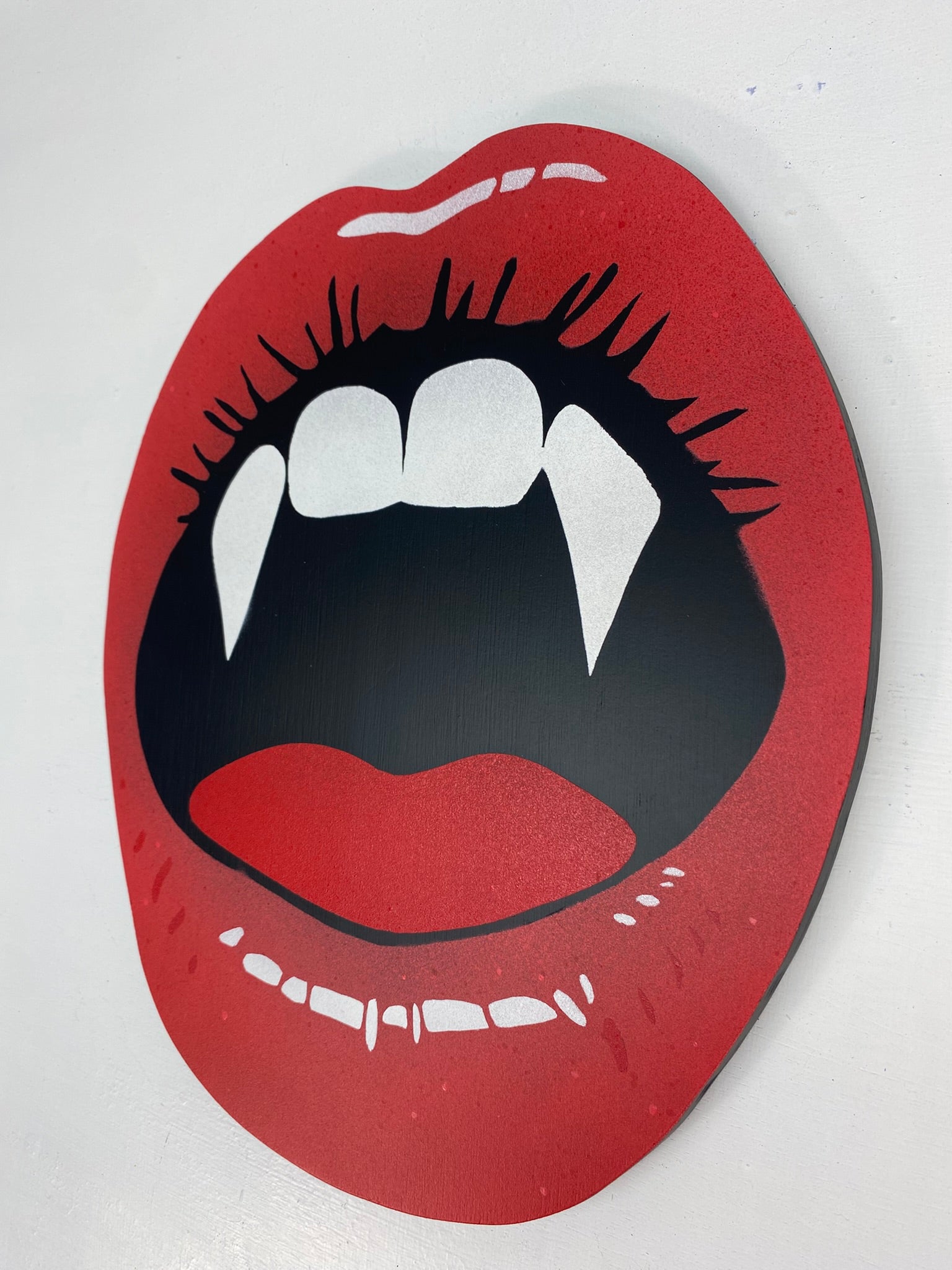 Hand made shaped spray-painting of red lips with vampire fangs on a white background.