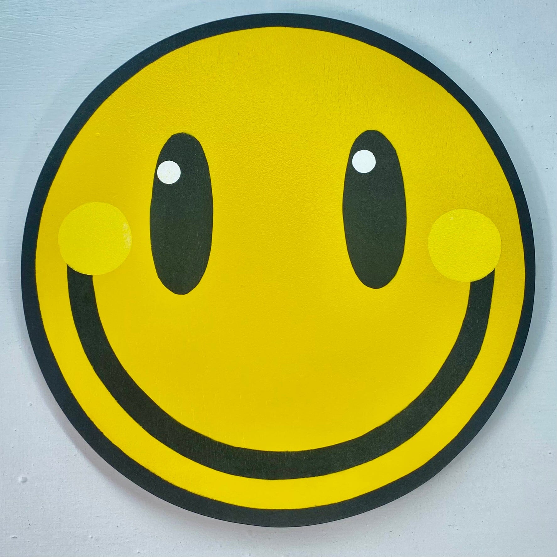 A square close up photo of a big yellow smiley spray-painting on a white painted wall.