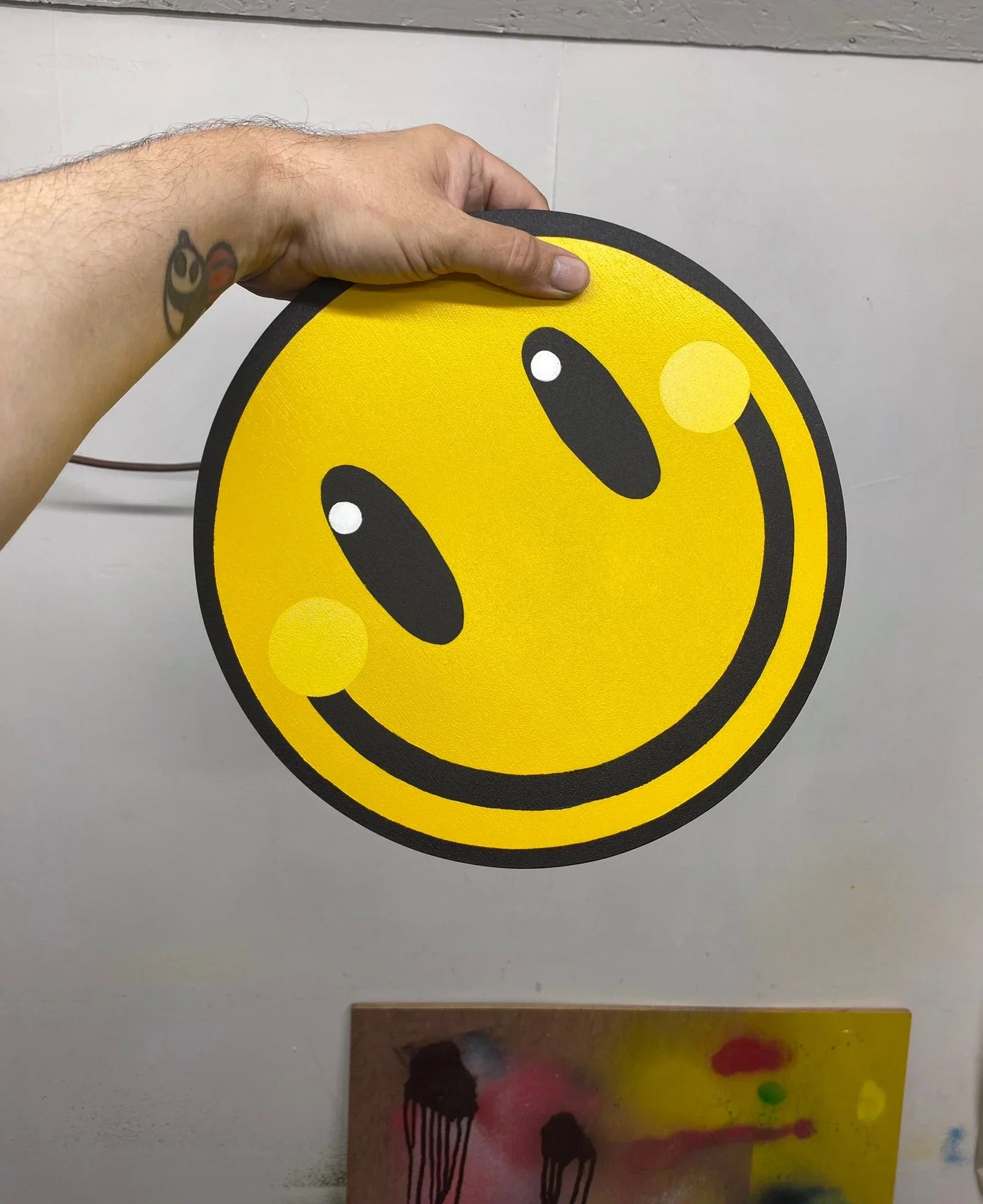 Hand holding a spray painted yellow smiley face wall art piece with black outline from WallShapes Shop.