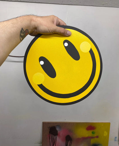 Hand holding a spray painted yellow smiley face wall art piece with black outline from WallShapes Shop.