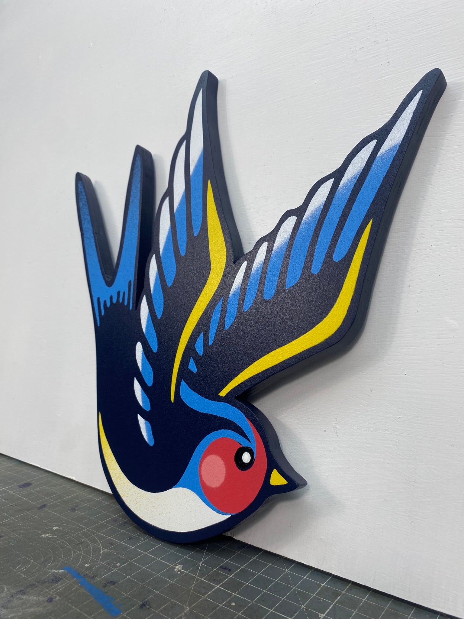 Tattoo inspired, colourful spray painting, painted on a hand-shaped wooden panel displayed on a stencil cutting mat, with a white wall.