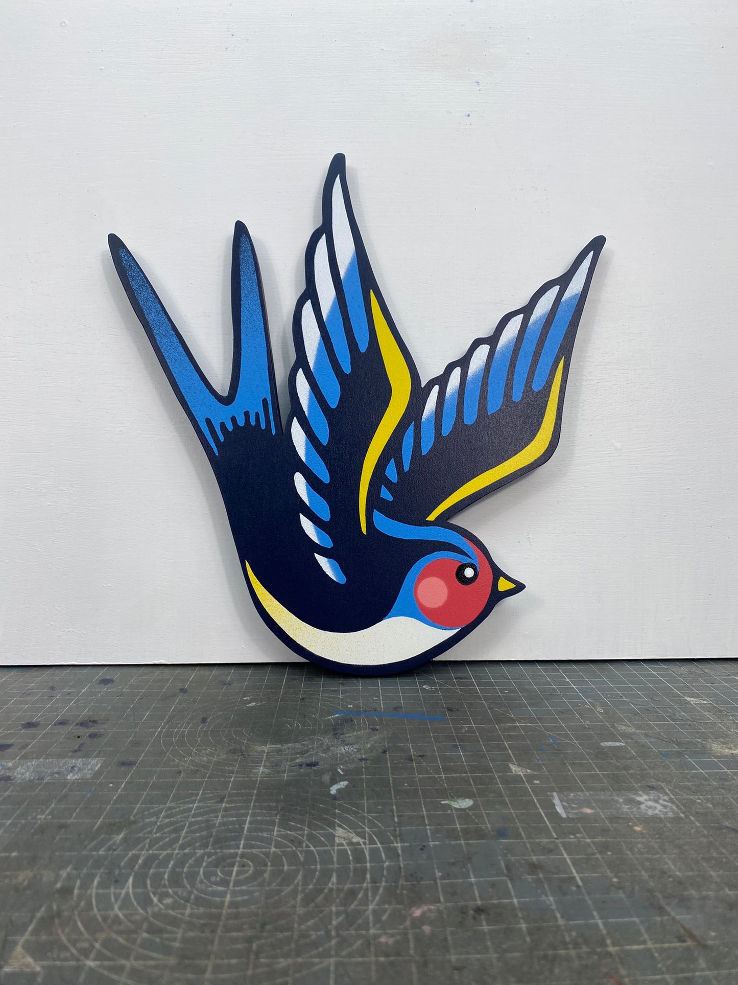 Tattoo inspired, colourful spray painting, painted on a hand-shaped wooden panel displayed on a stencil cutting mat, with a white wall.