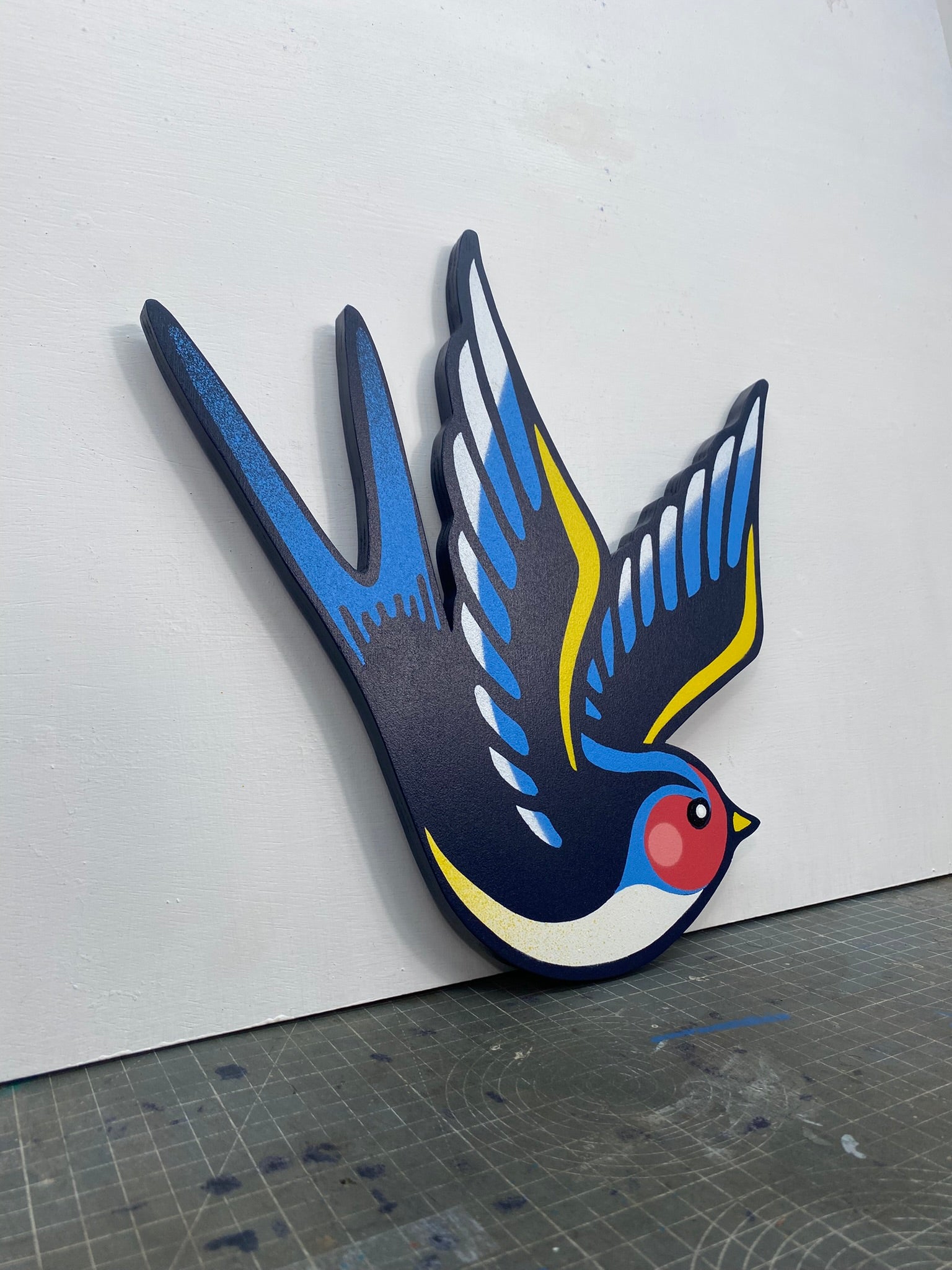 Tattoo inspired, colourful spray painting, painted on a hand-shaped wooden panel displayed on a stencil cutting mat, with a white wall.