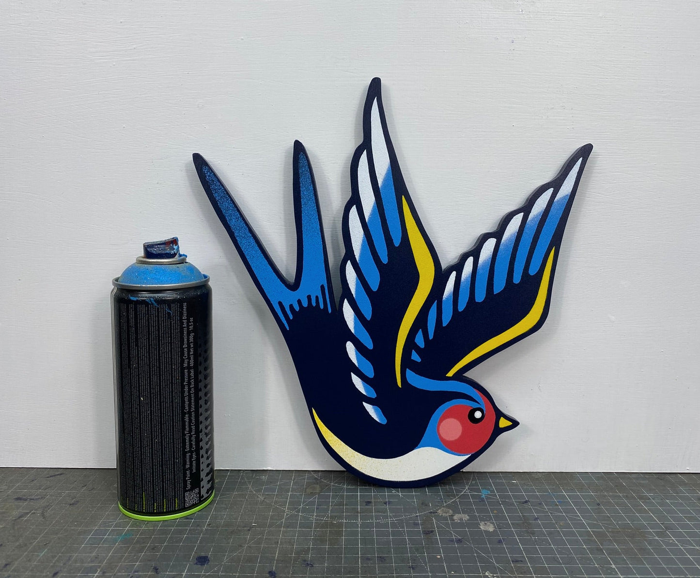 Tattoo inspired, colourful spray painting, painted on a hand-shaped wooden panel displayed on a stencil cutting mat, with a white wall. Alongside a spray can for size comparison.
