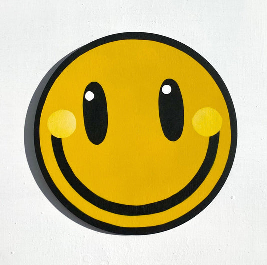 Smiley wooden wall art. Yellow happy face with with White highlights in black eyes and wide black smile, displayed on a White wall.