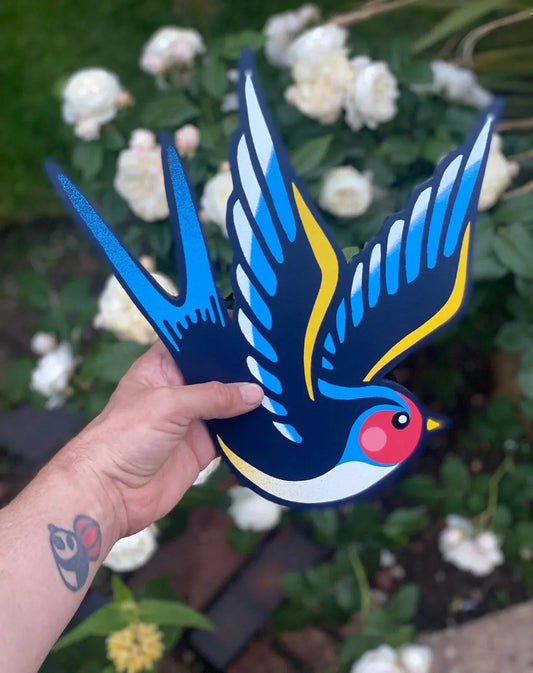 Hand holding colorful spray-painted Swallow bird wall art by WallShapes, with garden flowers background.