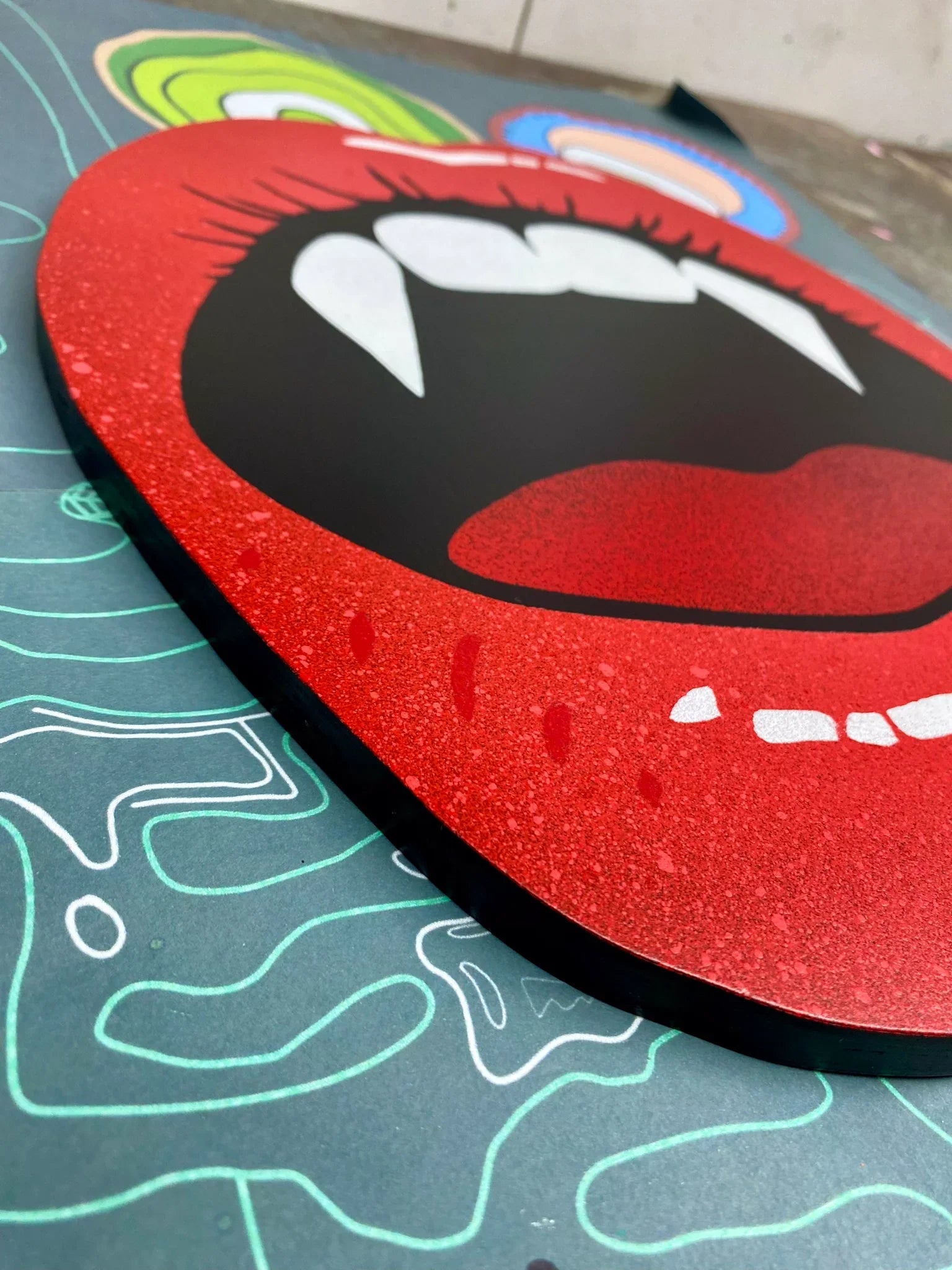 Handmade spray-painted wall art of red shiny lips with sharp white fangs displayed on an abstract background, from WallShapes Shop.