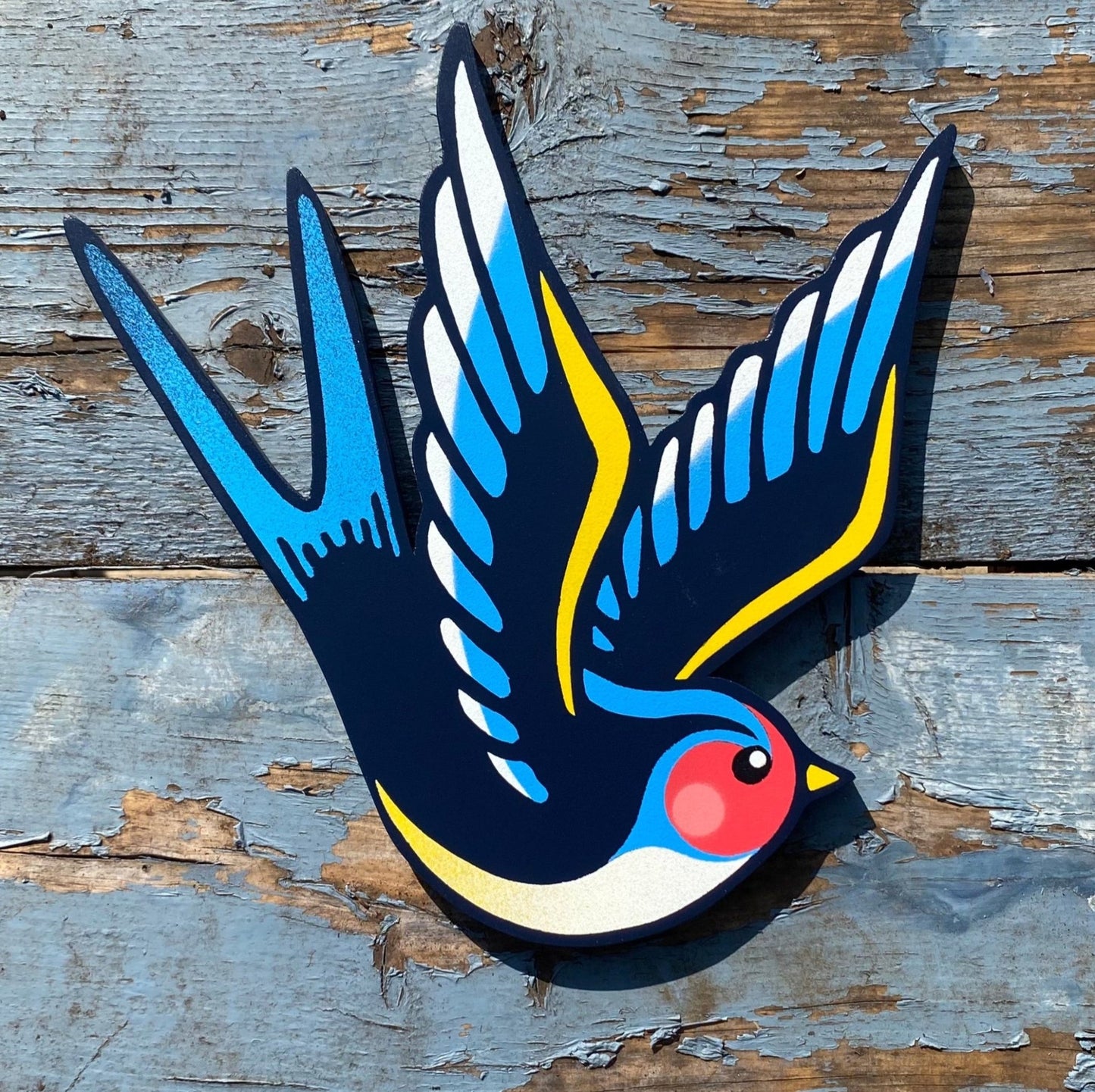 Hand-cut plywood swallow wall art painted in blue, yellow, and black with red cheek detail, inspired by vintage tattoo flash style.