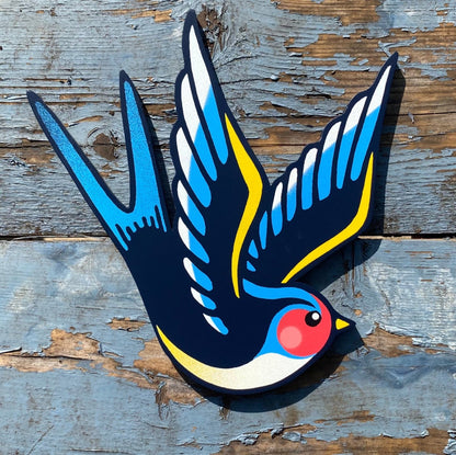 Hand-cut plywood swallow wall art painted in blue, yellow, and black with red cheek detail, inspired by vintage tattoo flash style.